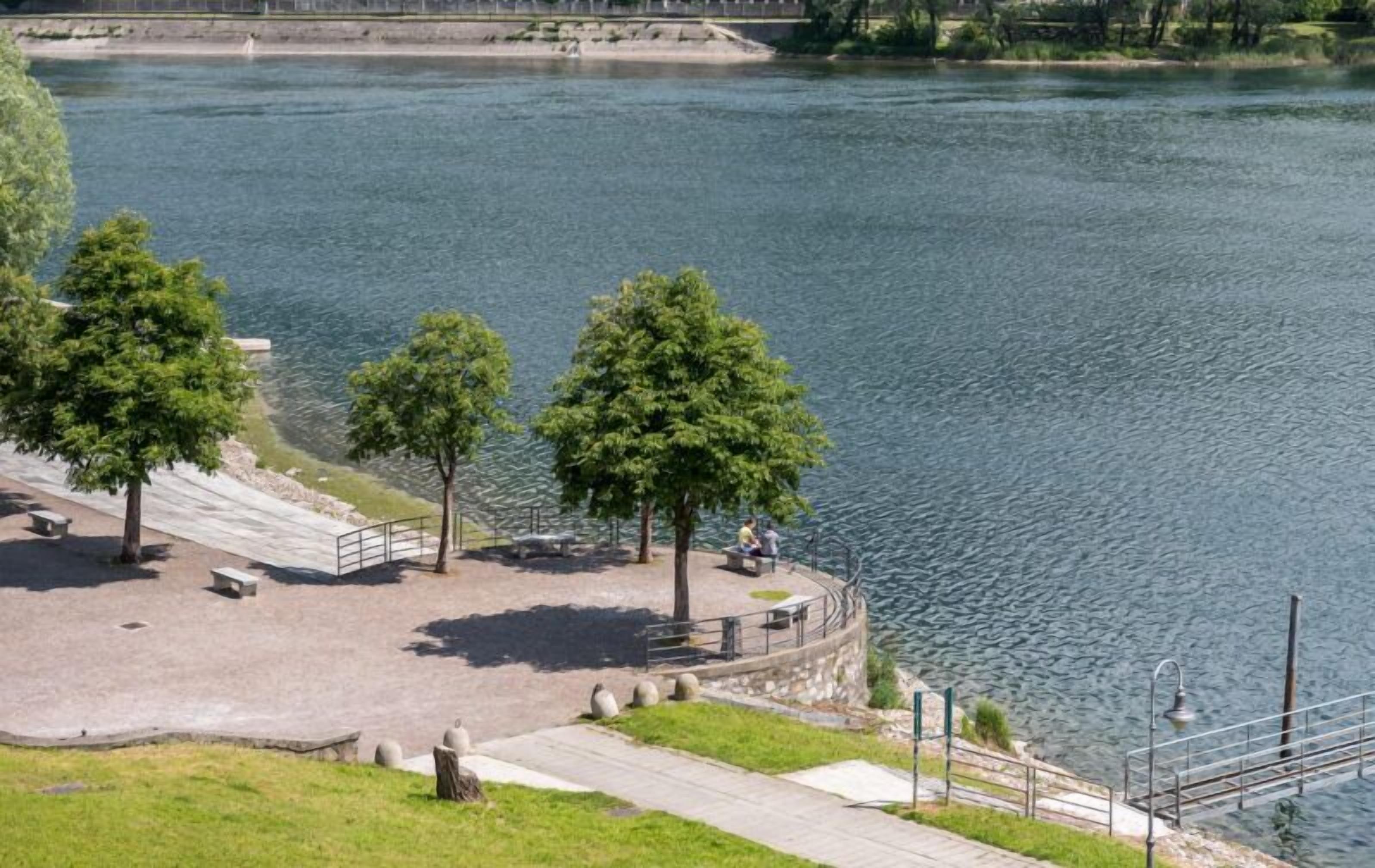 Um parque à beira-rio com bancos, árvores e uma trilha para caminhada.