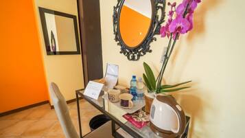 Un mostrador de recepción de hotel con un espejo, un jarrón con flores moradas, una botella de agua, una taza y un cartel.