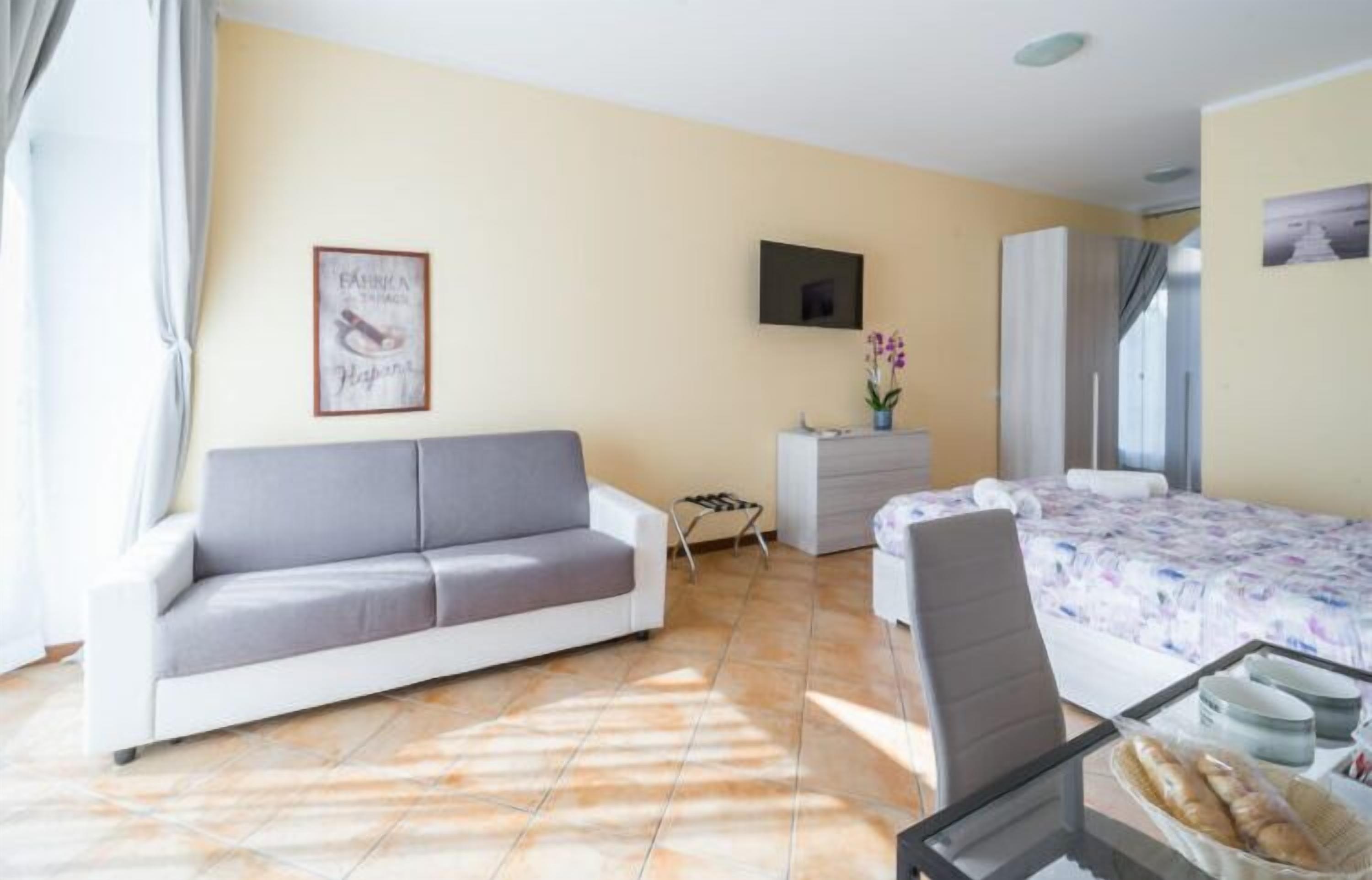 Una habitación con sofá, cama, escritorio con silla, televisor y una pequeña mesa con alimentos.