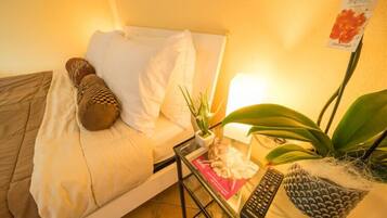 Una cama con almohadas, un control remoto, un libro y una planta sobre una mesita de noche.