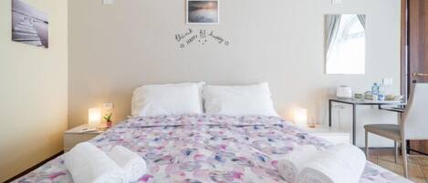 Habitación de hotel con una cama, dos mesitas de noche, un escritorio y una silla. Hay toallas sobre la cama y un cuadro en la pared.