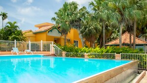 Ein Swimmingpool mit weißem Zaun, umgeben von Palmen und einem gelben Gebäude mit einem Ziegeldach.