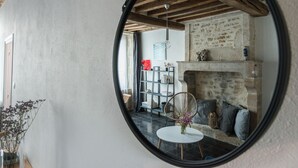 Un miroir rond reflète un salon avec une cheminée en pierre, une bibliothèque et une table ronde sur laquelle est posé un vase.