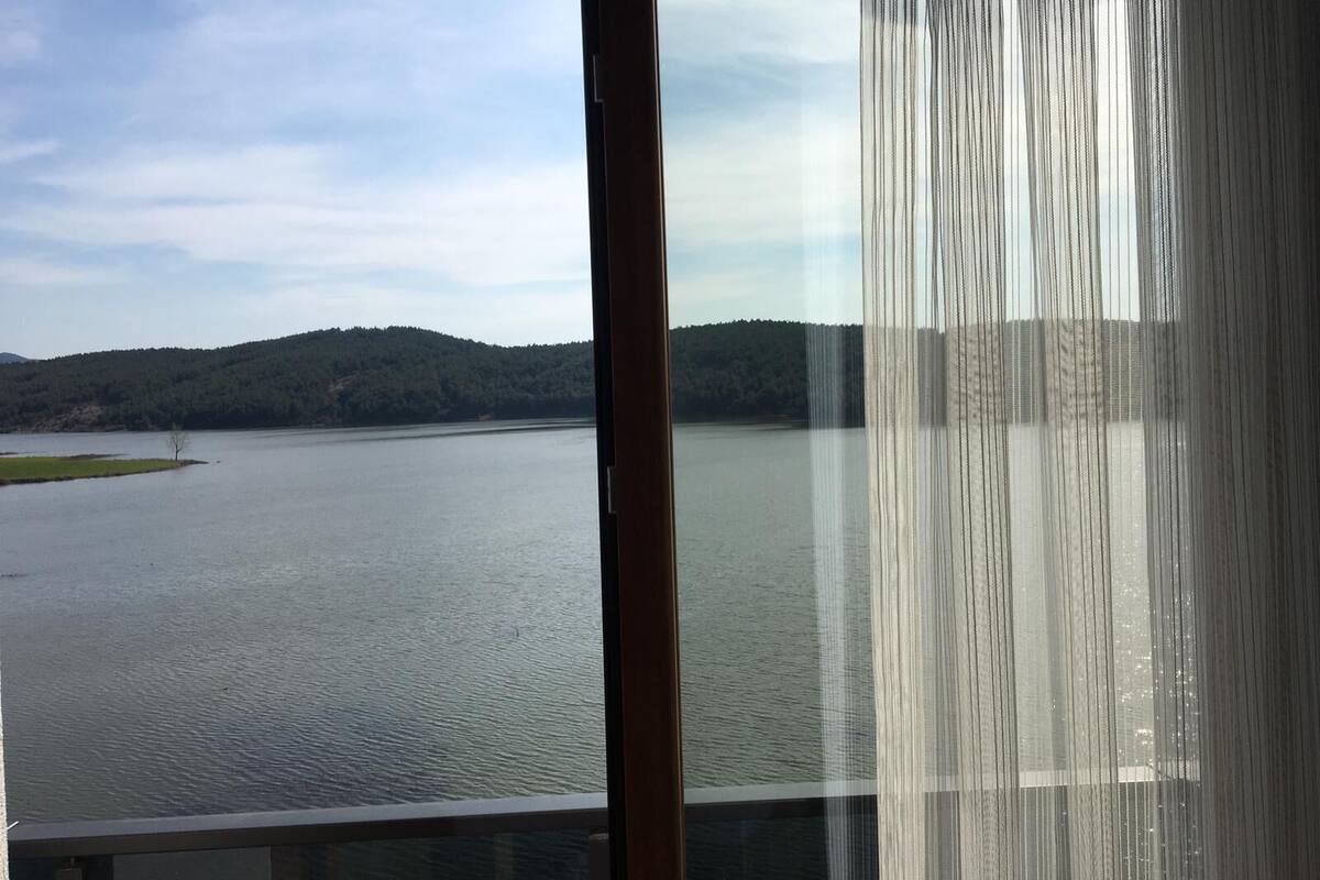 A view of a river with a forested hill in the background, seen through a window with sheer curtains.