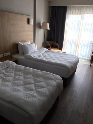 Une chambre d’hôtel avec deux lits, une tête de lit en bois, une chaise, une lampe et une vue sur l’extérieur à travers des rideaux transparents.