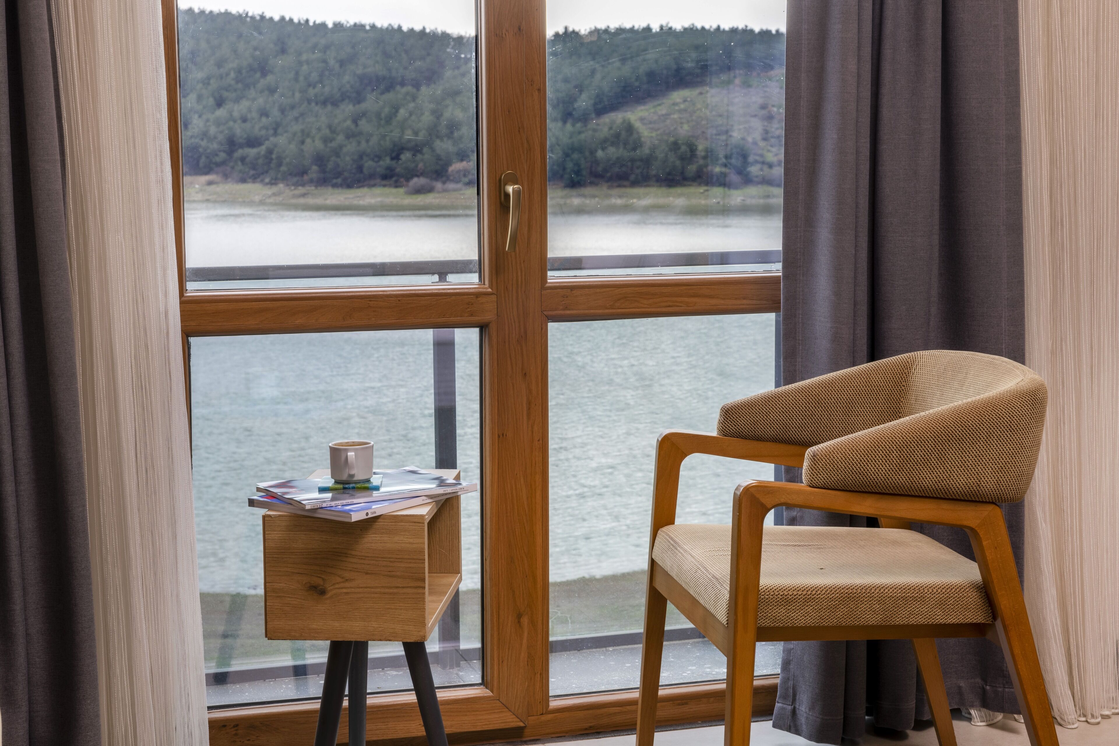 A room with a wooden chair, a small table with a cup and magazines, and a large window with a view of a forest and water.