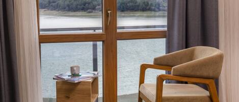 A room with a wooden chair, a small table with a cup and magazines, and a large window with a view of a forest and water.