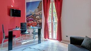 Une pièce avec un mur rouge, un téléviseur à écran plat, une table en verre avec des chaises, une grande affiche de voitures de dessins animés et un balcon avec des rideaux rouges.