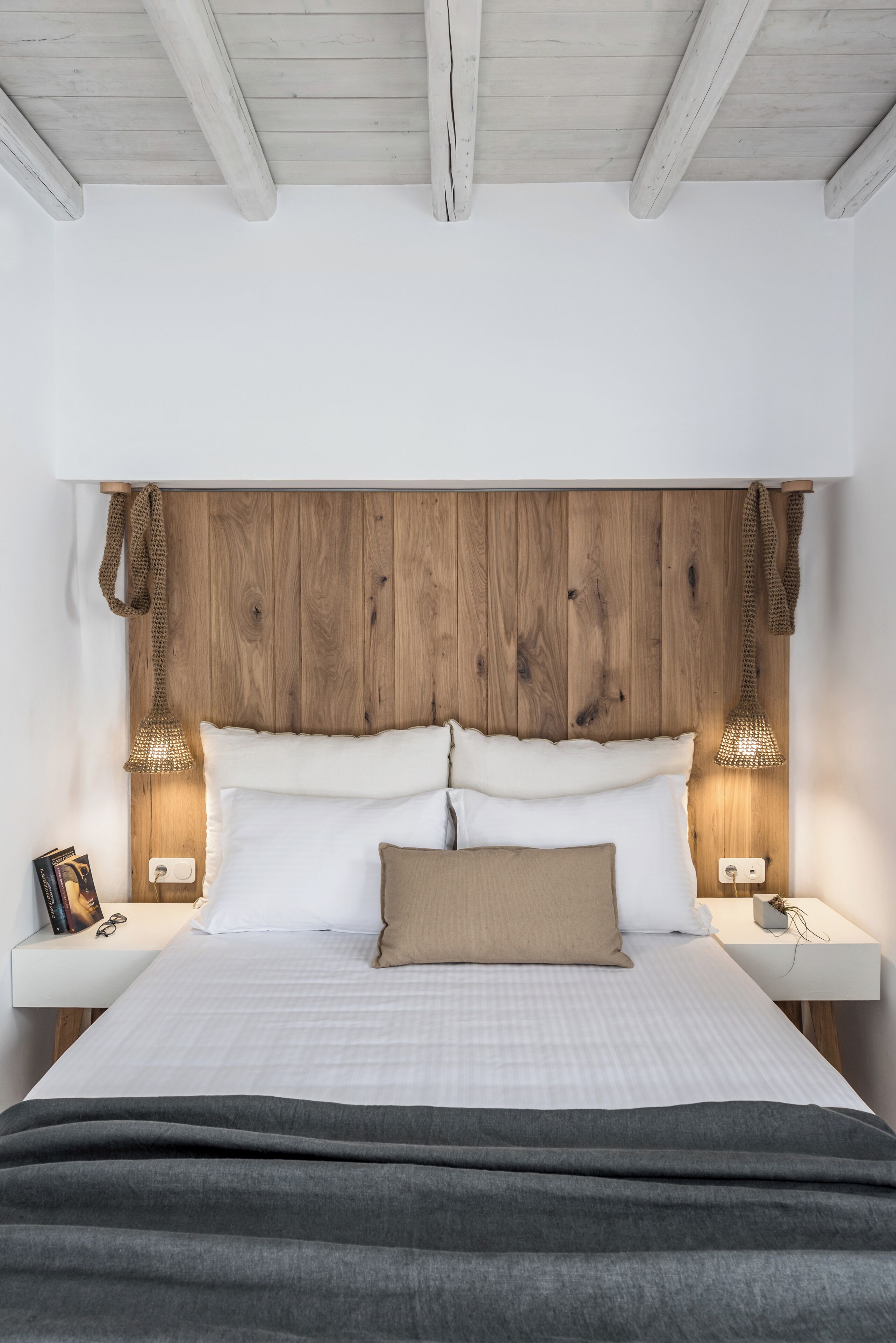 A bed with a wooden headboard, two bedside lamps, a bedside table with a book and glasses, and a blanket.