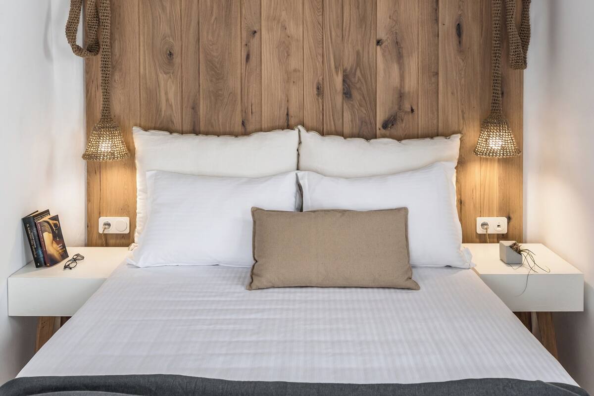 Une chambre à coucher avec une tête de lit en bois, une literie blanche, deux lampes de chevet et une table de chevet sur laquelle se trouvent un livre et des lunettes.