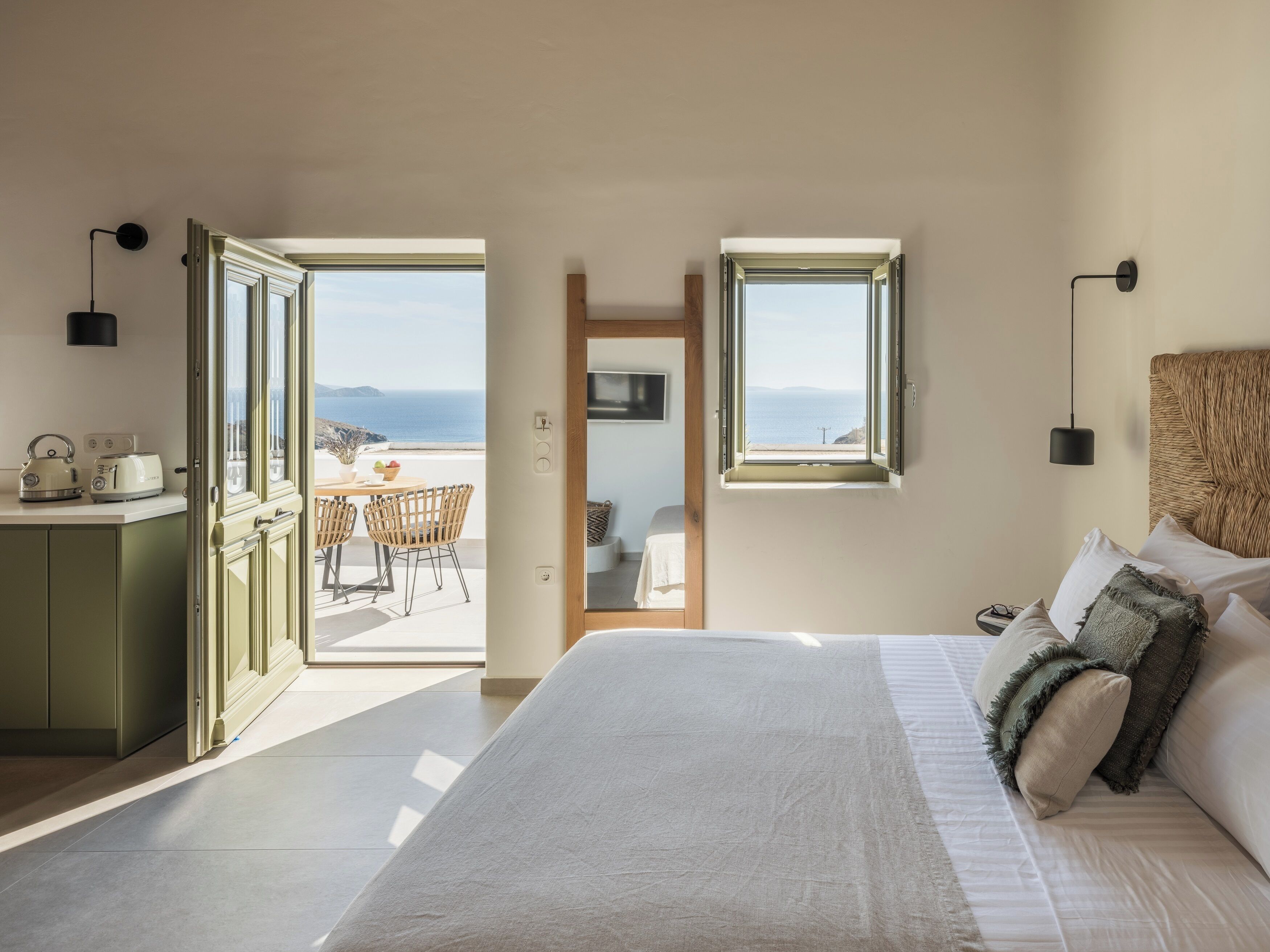 A bedroom with a large bed, a view of the sea through a window, and a balcony with a table and chairs.