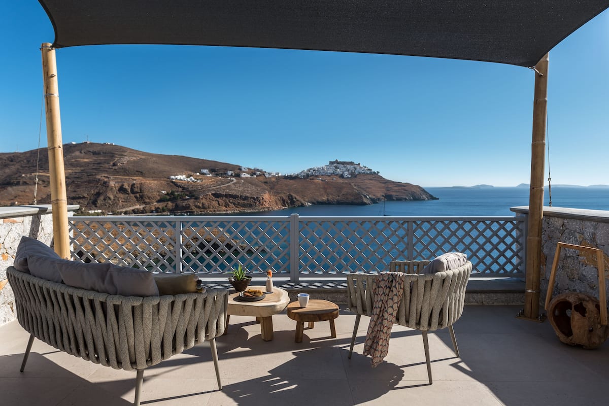 Une terrasse meublée de mobilier en osier, une petite table agrémentée d’une plante, et une vue sur un paysage côtier avec une colline et la mer.