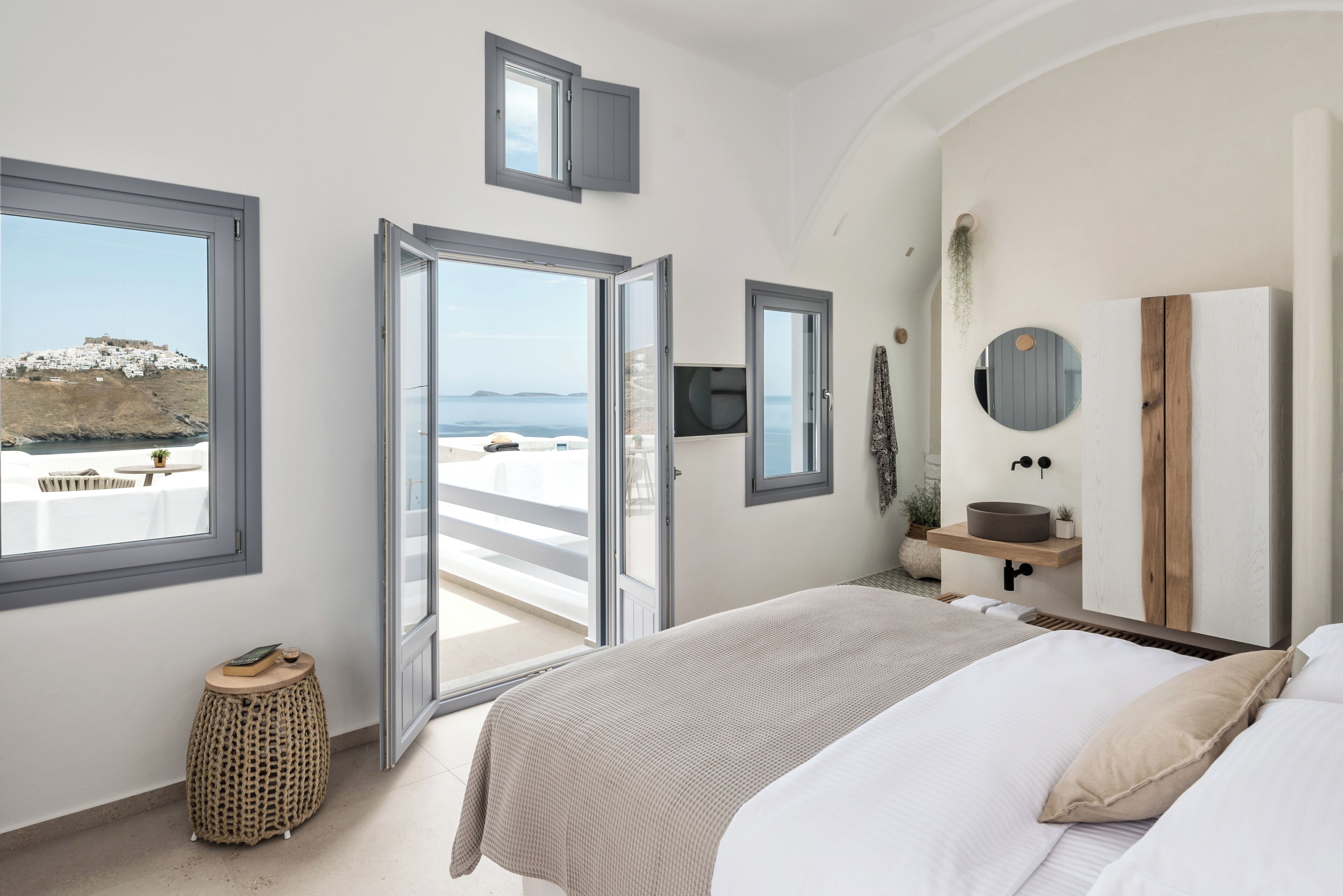 A bedroom with a bed, a bedside table, a mirror, a window with a view of a beach, and a door leading to an outdoor balcony.