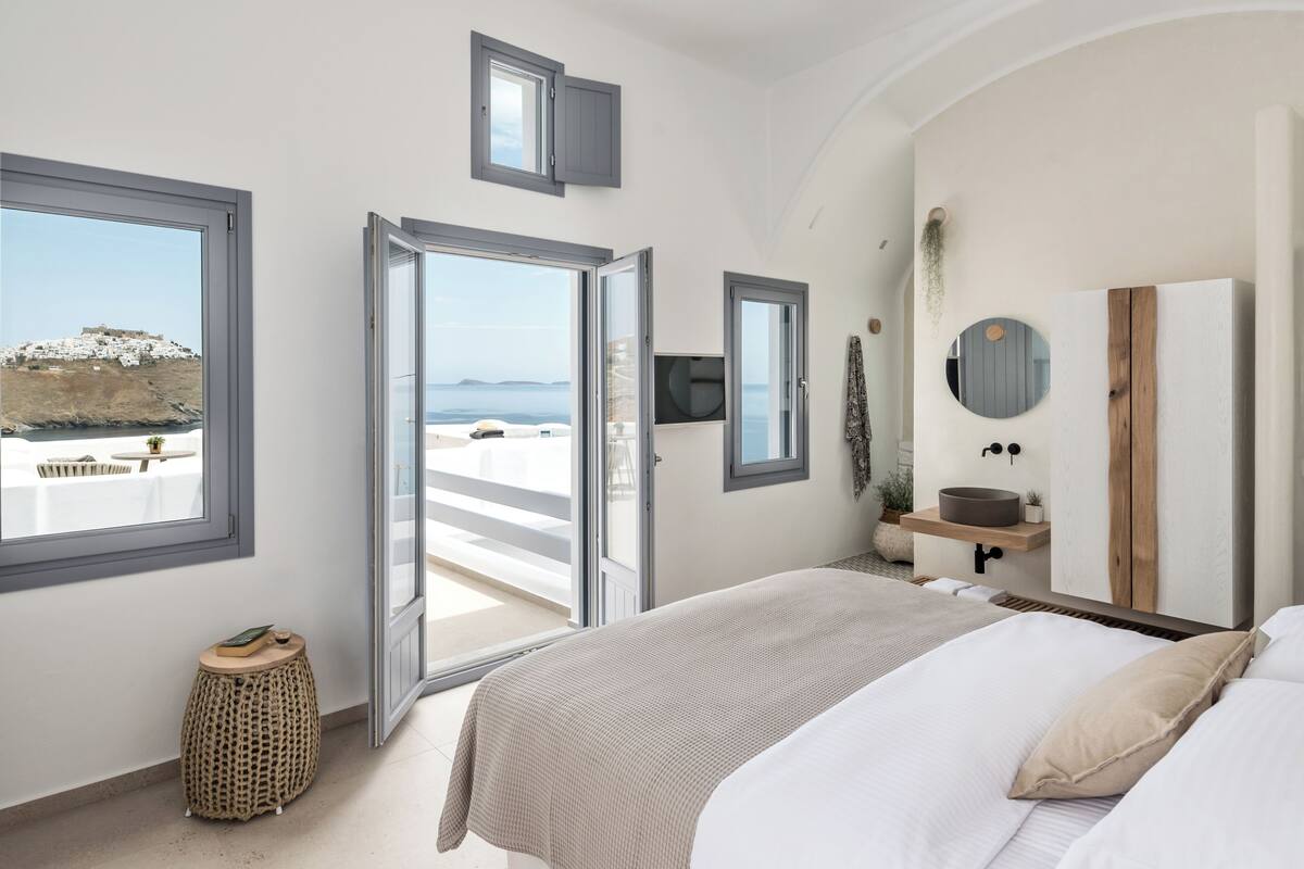 Une chambre dotée d’un grand lit, d’une table de chevet, d’un miroir, d’une fenêtre donnant sur la mer et d’une porte menant à un balcon extérieur.