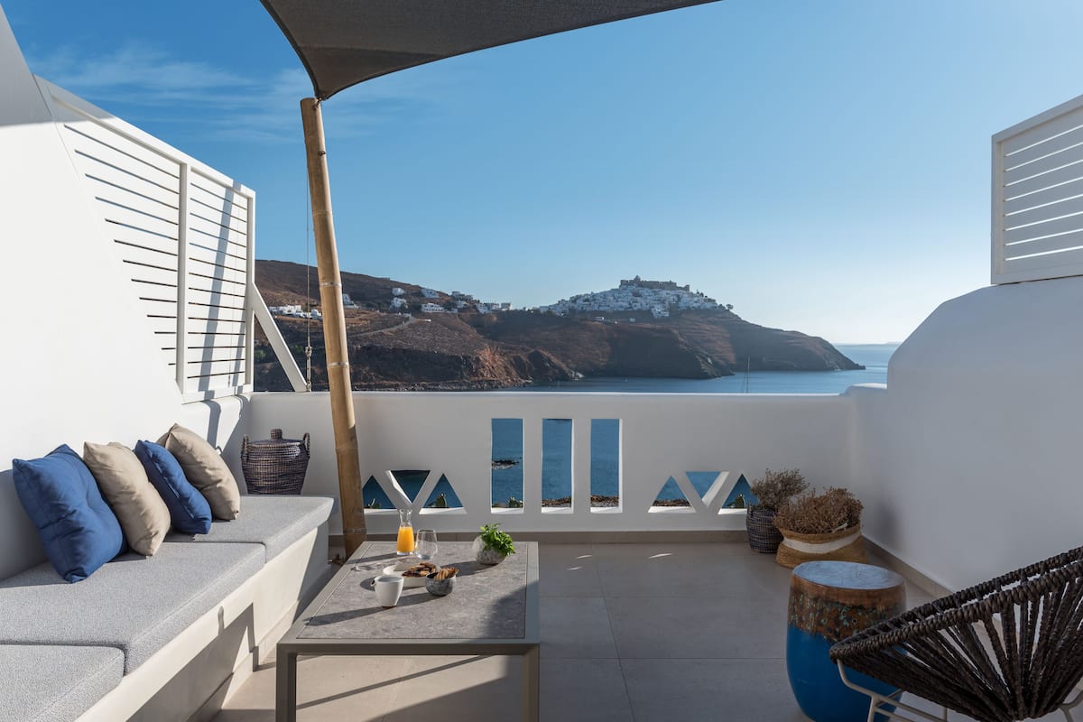Un balcon avec un canapé blanc, des coussins, une table sur laquelle se trouve un verre de jus d’orange, une chaise en osier et une vue sur une ville côtière aux bâtiments blancs.