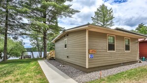 A small, single-story cabin with a wooden exterior, a concrete pathway leading to the entrance, and a bench near the water.
