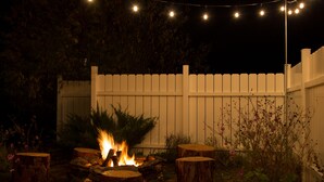 Escena nocturna al aire libre con un pozo de fuego rodeado de taburetes de madera, una valla de piquetes blancos y luces colgantes.