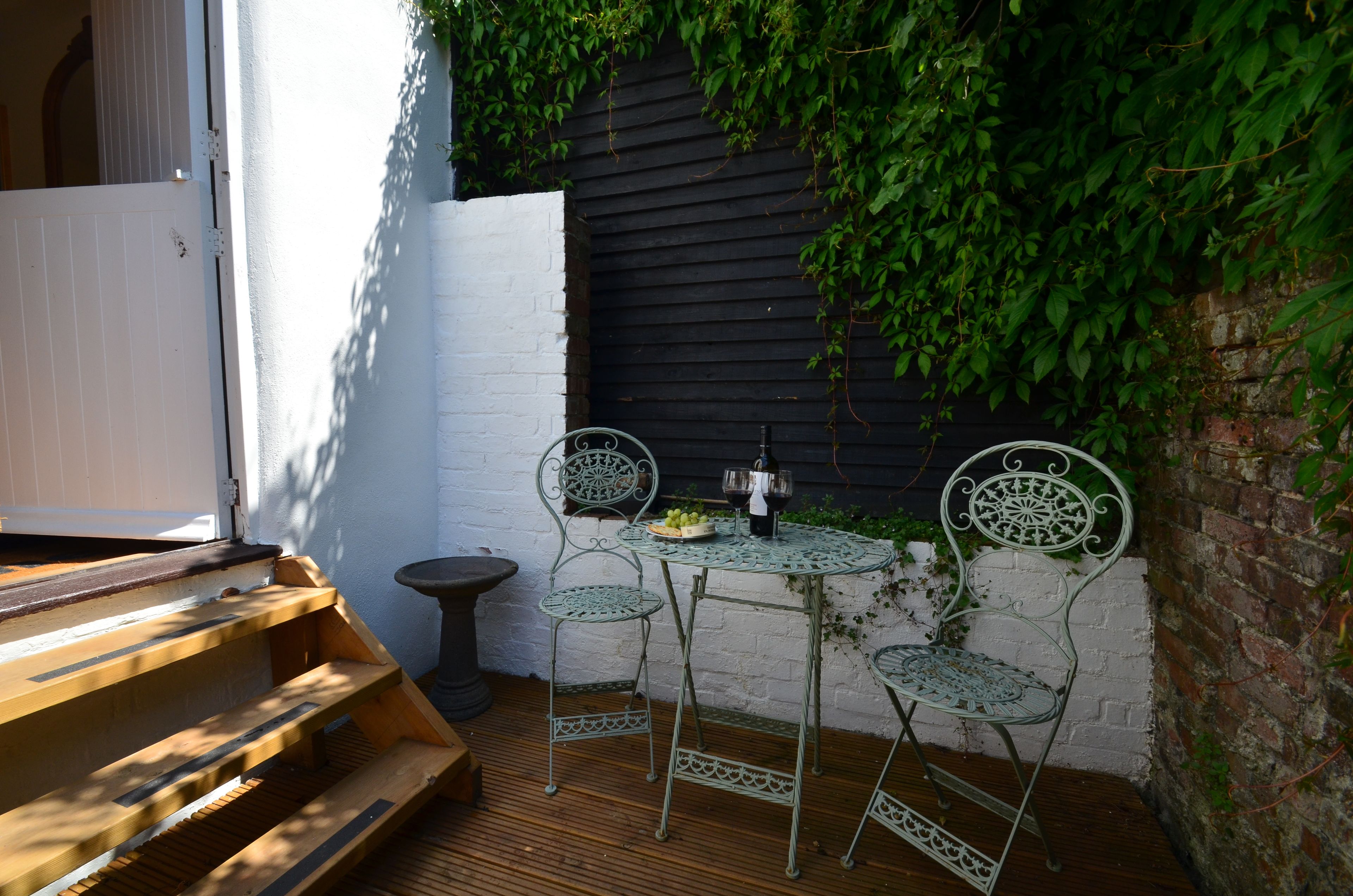 A small outdoor patio with a wooden bench, two metal chairs, a small round table, and a bottle of wine.