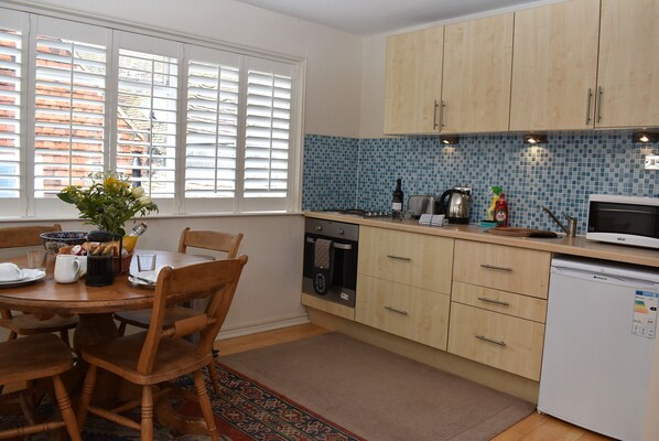 A kitchen with wooden cabinets, a tiled backsplash, a dining table with chairs, and a window with blinds. - Studio 22 (Rye)