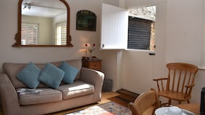A cozy living room with a sofa, a wooden chair, a dining table with a plate and a cup, a mirror, and a door with shutters.