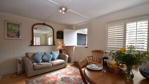 A cozy living room with a sofa, a dining table with a vase of flowers, and a large window with blinds.