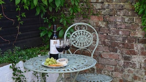 A table with a bottle of wine, two glasses, a plate of grapes, and a small container of cheese, set against a brick wall with greenery.