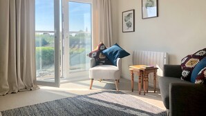 Une pièce dotée d’une porte-fenêtre coulissante donnant sur un balcon, d’un fauteuil blanc agrémenté d’un coussin coloré, d’un canapé foncé, d’un tapis à motifs et de deux reproductions botaniques encadrées accrochées au mur.