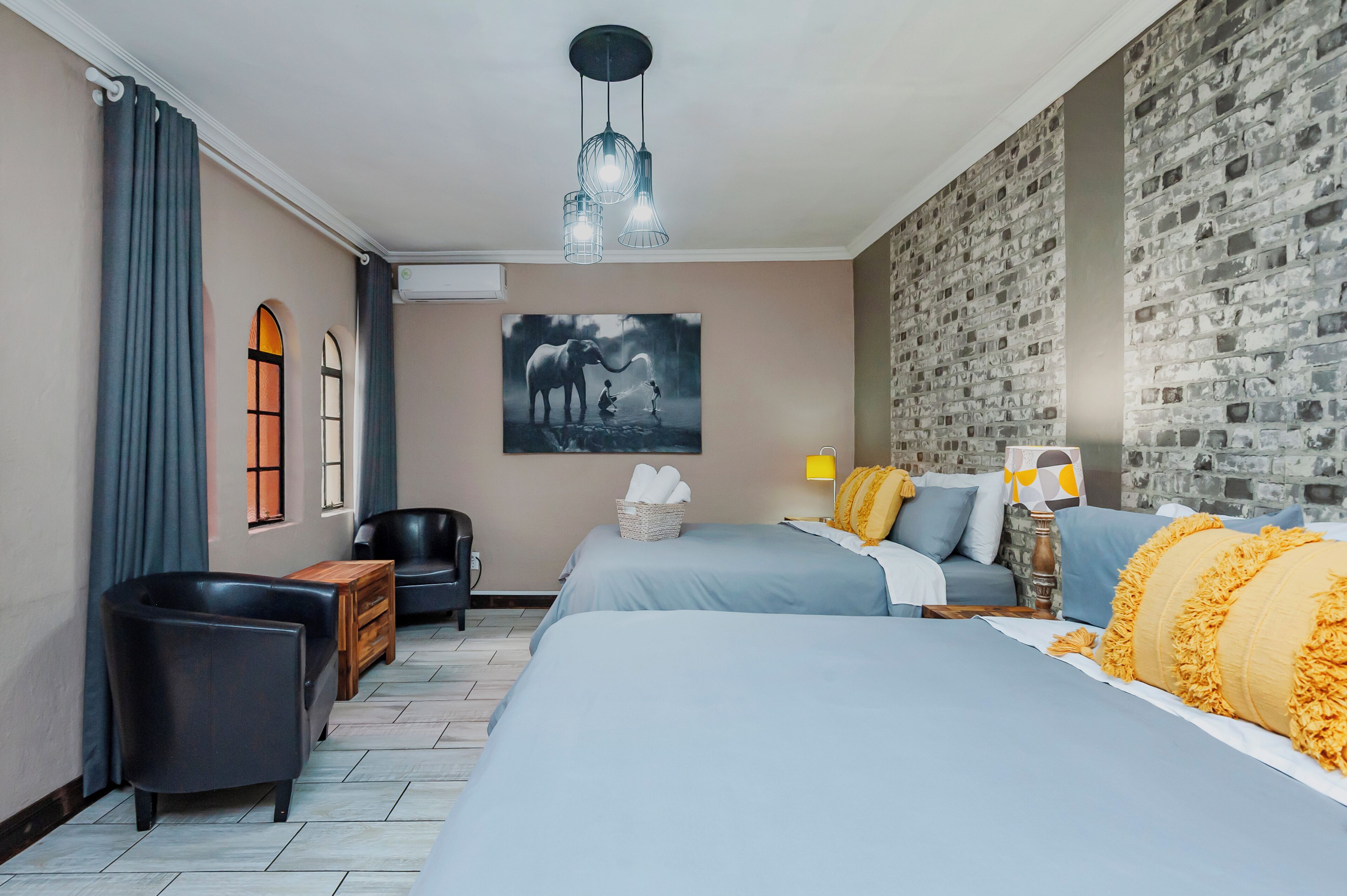 A hotel room with two beds, a large window with curtains, a wall-mounted air conditioner, a black leather chair, a wooden side table, a lamp, and a wall art of an elephant.