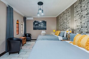 Habitación de hotel con dos camas, un ventanal con cortinas, aire acondicionado en la pared, un sillón de cuero negro, una mesita de noche de madera, una lámpara y una obra de arte en la pared con un elefante.
