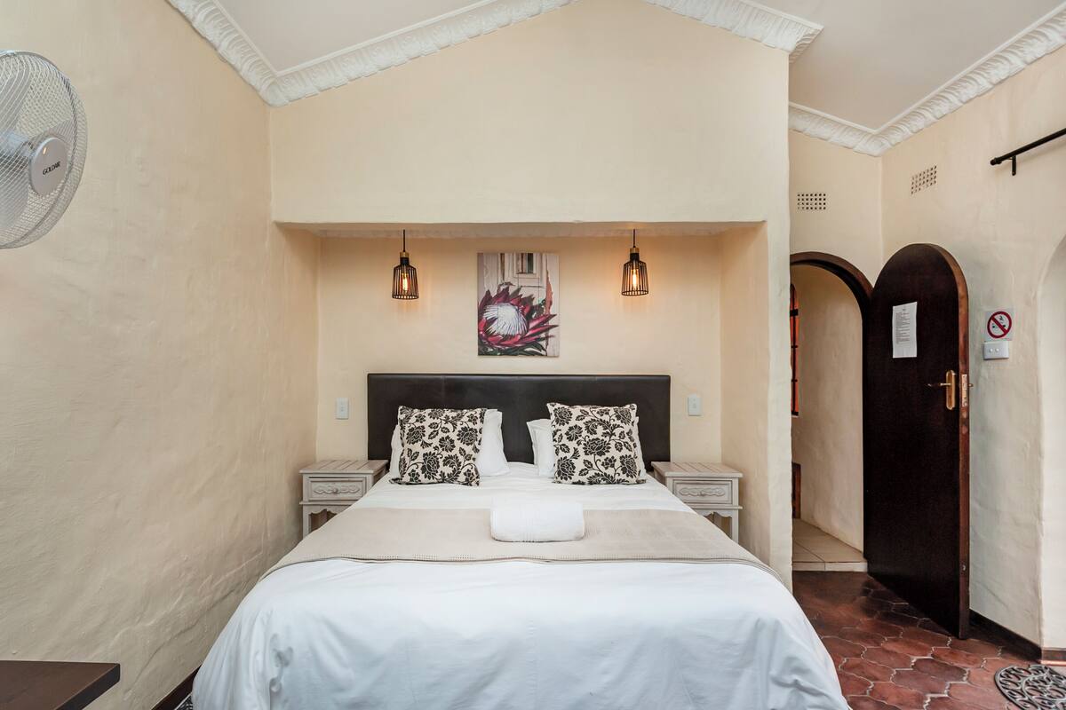 A hotel room with a bed, two bedside tables, a wall-mounted fan, and a door with a no-smoking sign.