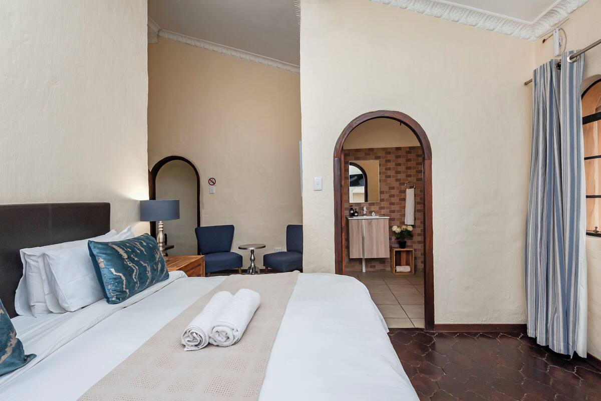 A hotel room with a large bed, two armchairs, a small table, and a bathroom visible through an archway.