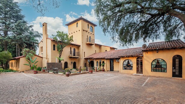 A courtyard with a cobblestone floor, a building with a tower, and surrounding greenery. - Airport Tower Lodge (Kempton Park)