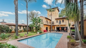 A pool area with a brick patio, palm trees, and a building with a tiled roof and arched windows. - Airport Tower Lodge (Kempton Park)