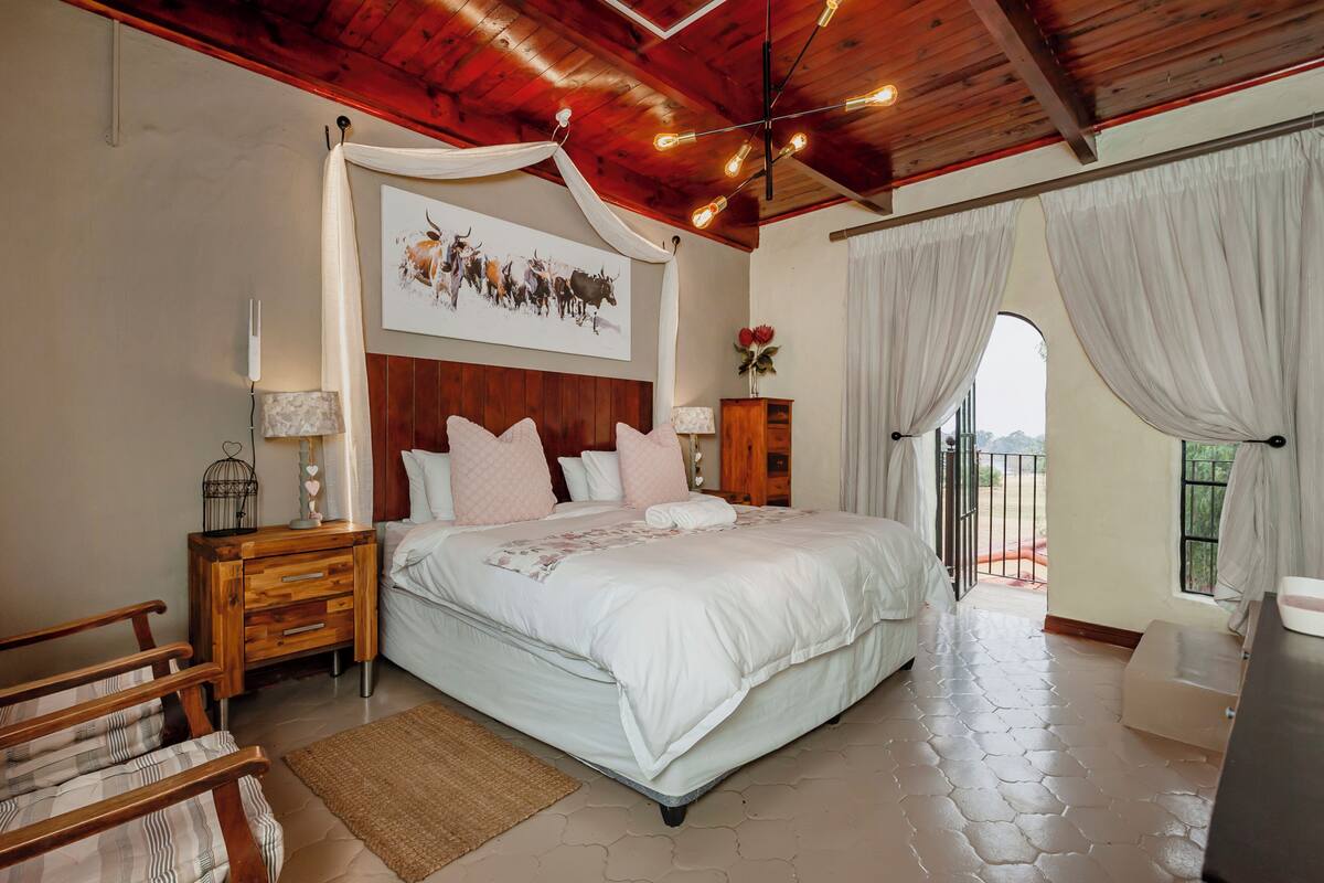 A bedroom with a large bed, wooden headboard, bedside tables, a nightstand, a lamp, a framed picture, and a view through a window with curtains.