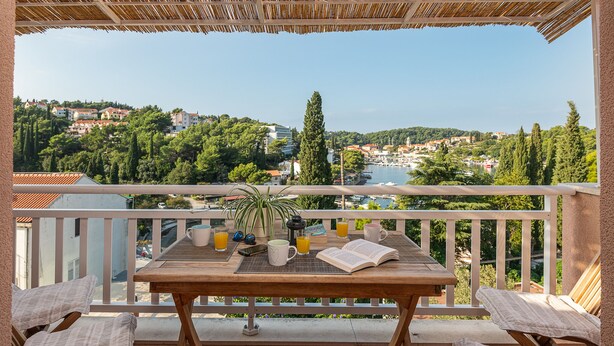 Ein Balkon mit Holztisch, zwei gepolsterten Stühlen, einem Blumentopf und einem Buch.