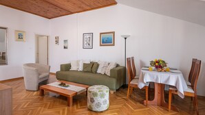 Ein Wohnzimmer mit Holzbalkendecke, einem grünen Sofa, einem runden Hocker, einem Esstisch mit Stühlen und einer Stehlampe.