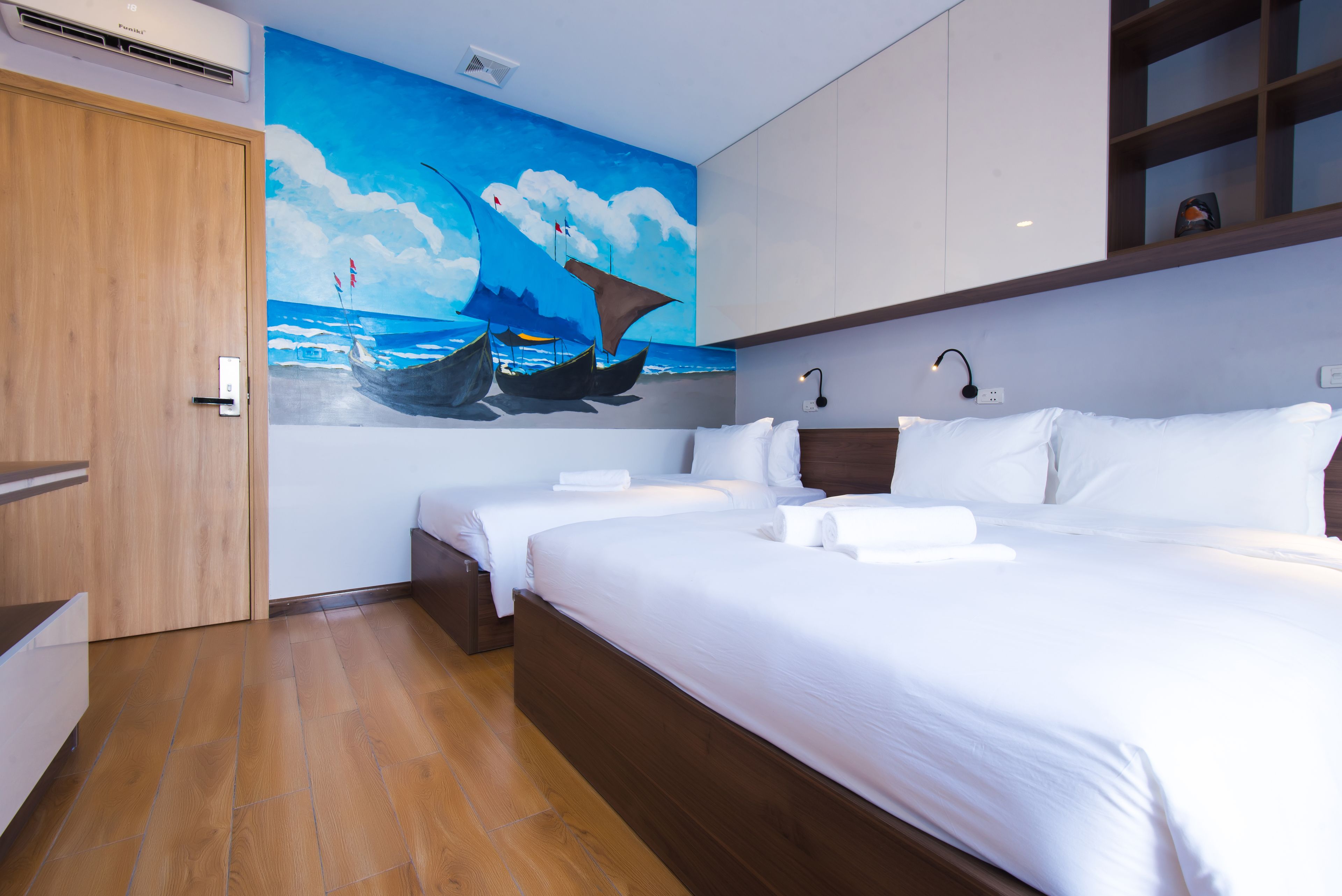 Une chambre d’hôtel avec deux lits, un plancher en bois, une fresque représentant des bateaux sur le mur et une étagère intégrée au-dessus des lits.