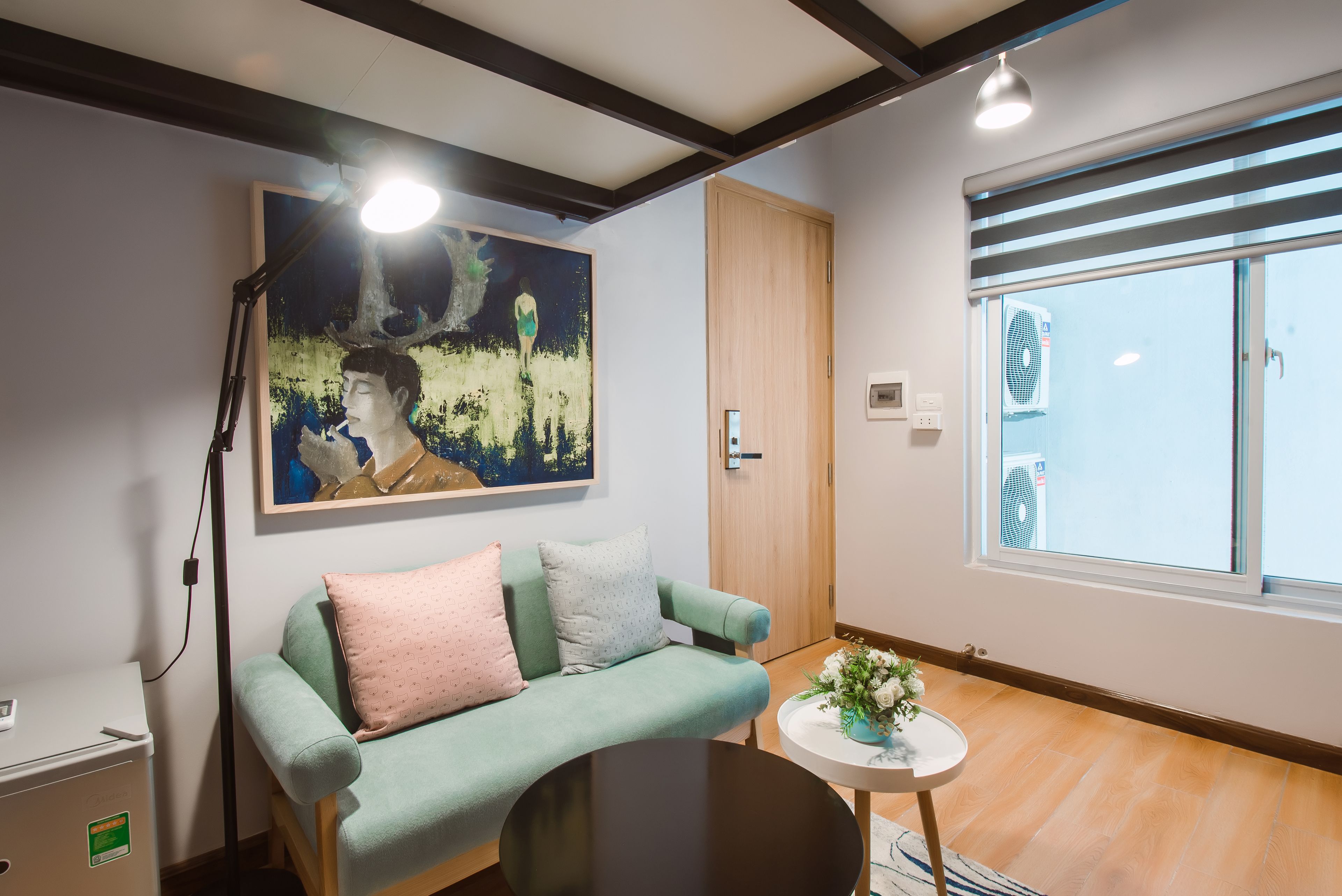 A modern living room with a teal sofa, a small round table with a vase, a painting on the wall, and a window with blinds.