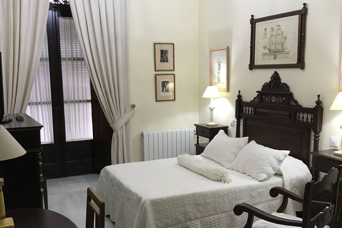 Une chambre d’hôtel avec un lit, un bureau, une chaise, un lustre et une fenêtre avec des rideaux.