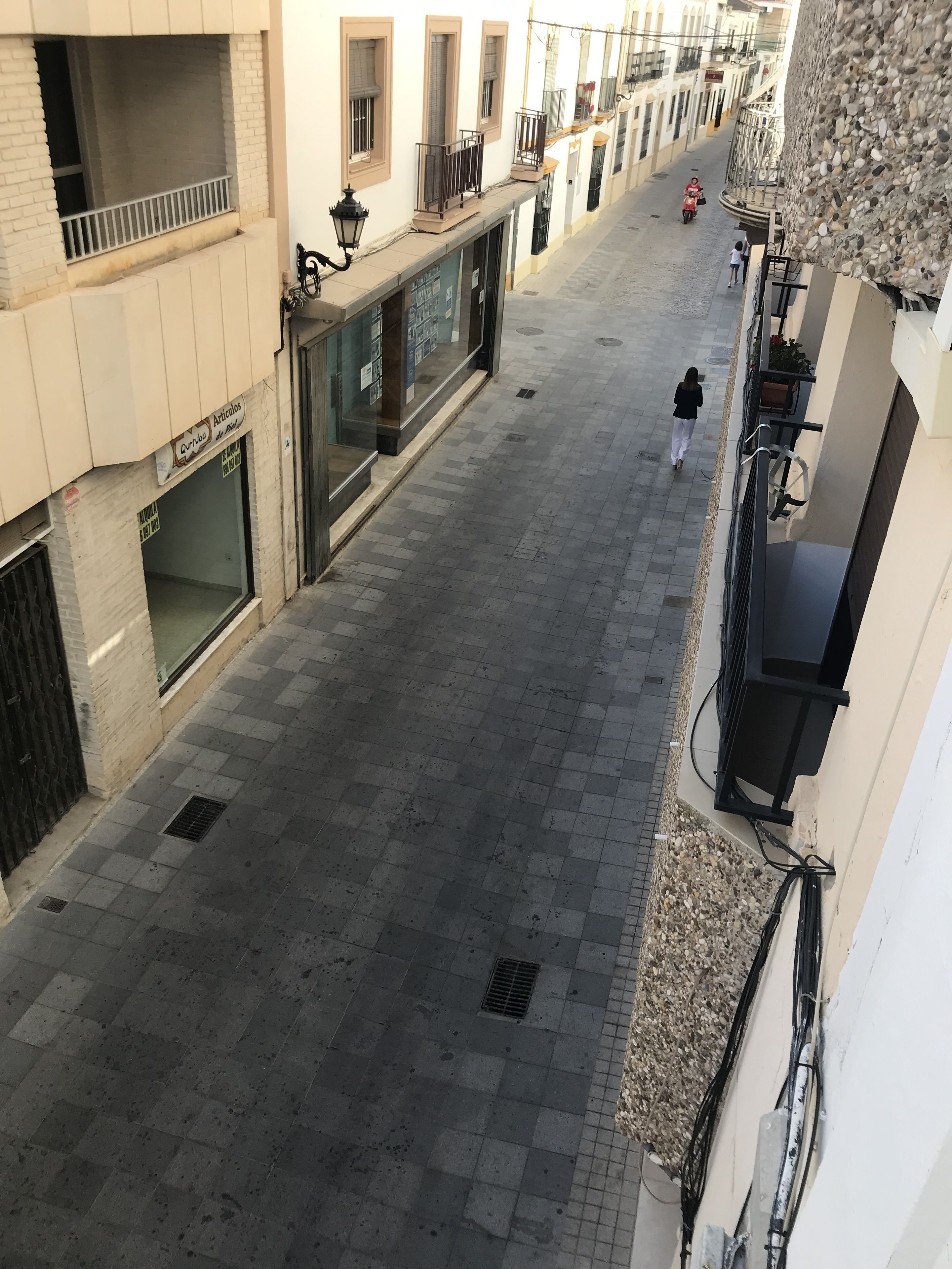 A narrow street with buildings on both sides, featuring balconies, windows, and a few pedestrians.