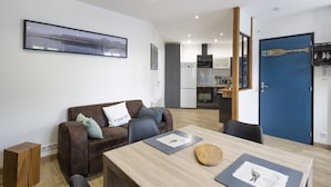 A modern living room with a brown leather sofa, a wooden coffee table, and a dining table with chairs. There is a kitchen area in the background with a blue door and a framed picture on the wall.