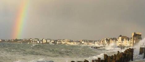 A coastal scene with a rainbow, waves crashing against a stone pier, and buildings in the background.