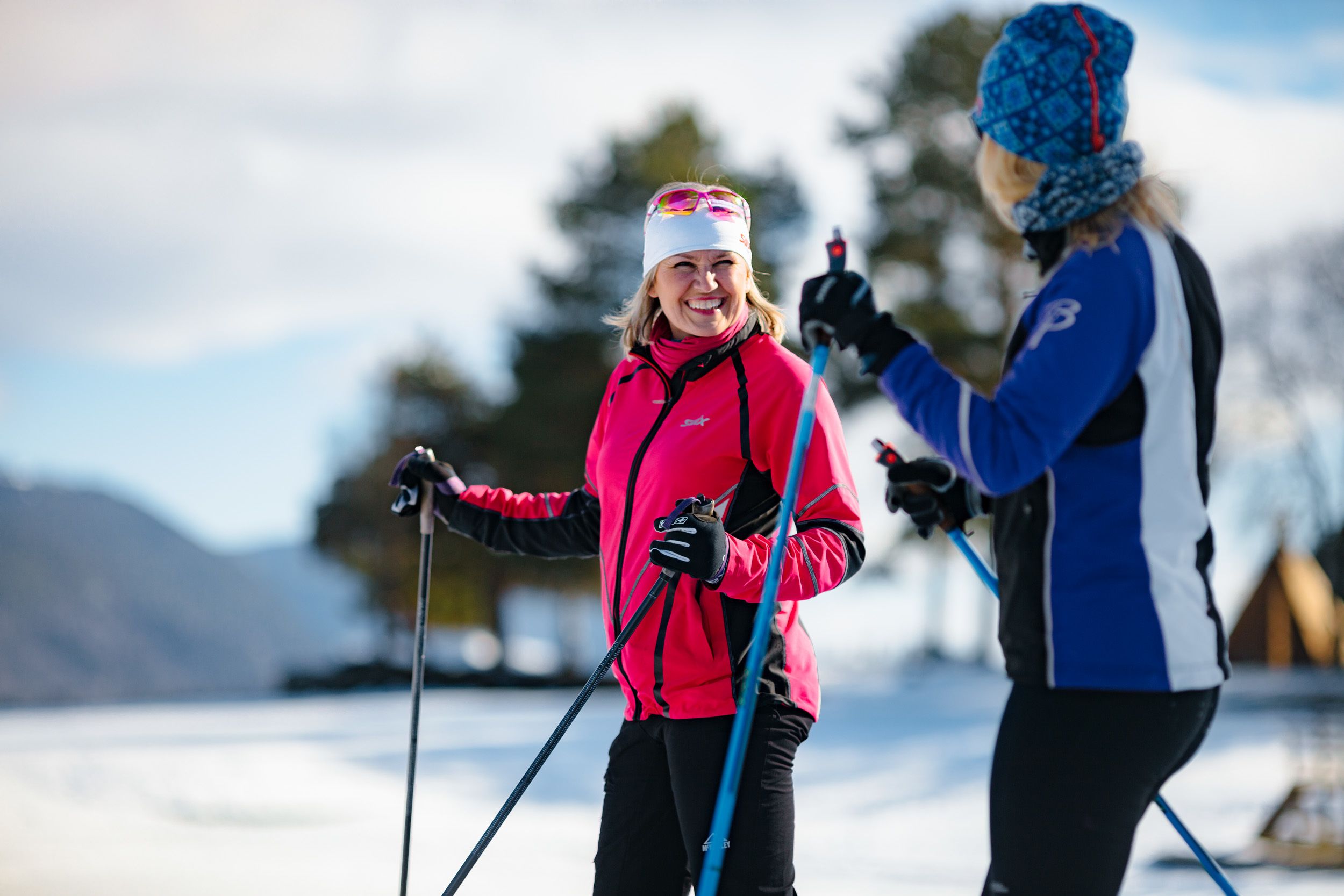 Two people skiing, one in a red jacket and the other in a blue jacket, with snow and trees in the background.