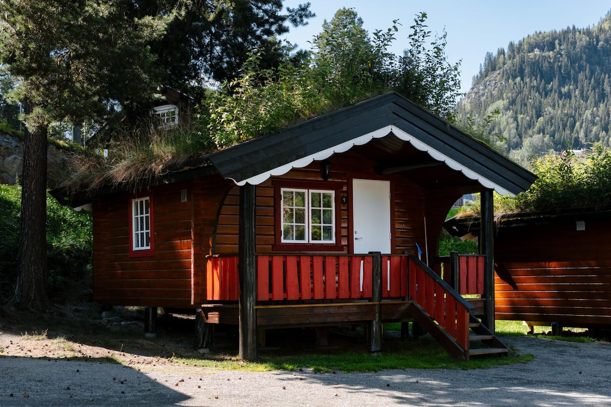 A small wooden cabin with a porch and a staircase, surrounded by trees and a mountain in the background.