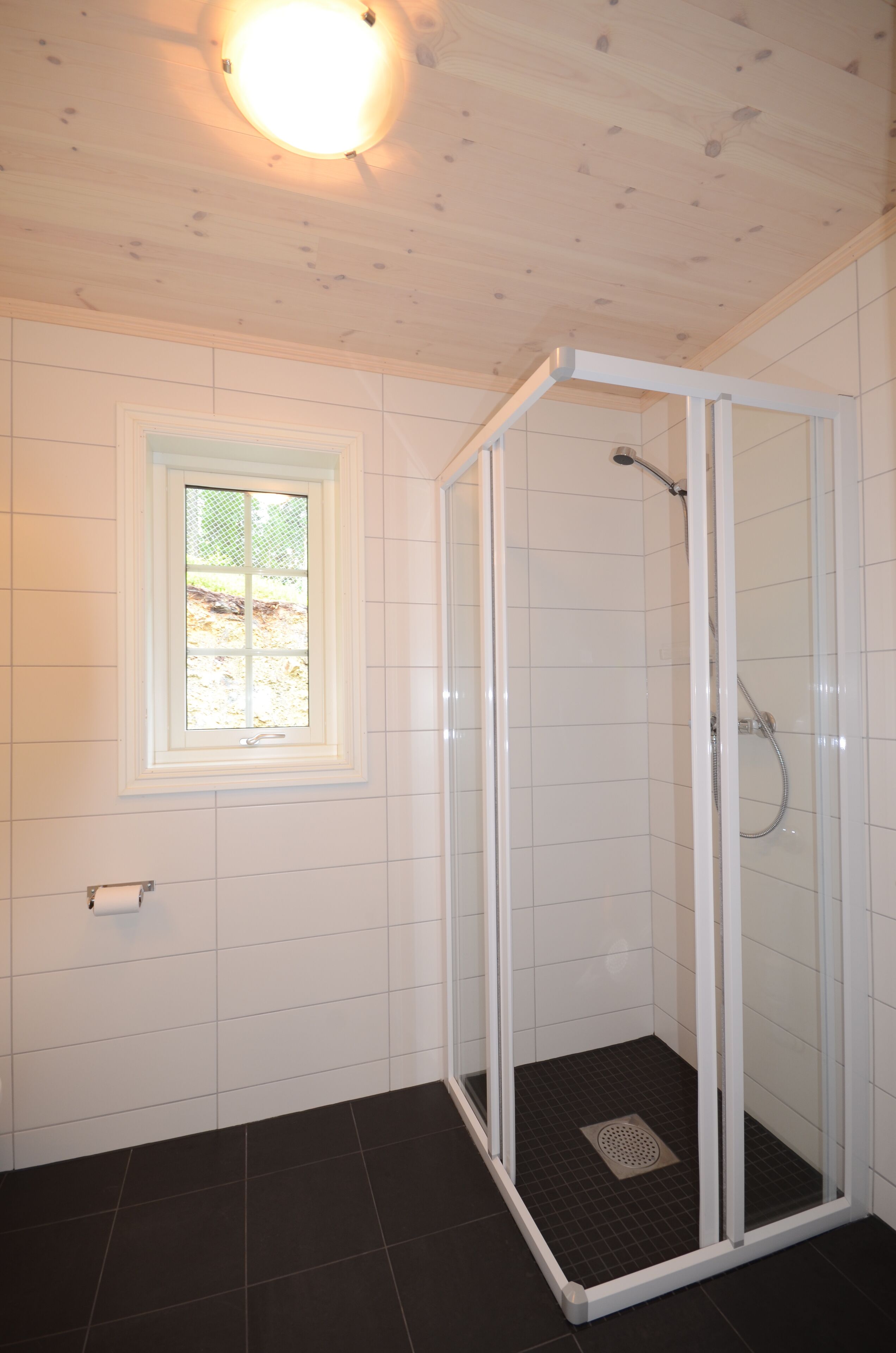 A shower area with a glass door, white tiled walls, a window, and a roll of toilet paper on the wall.