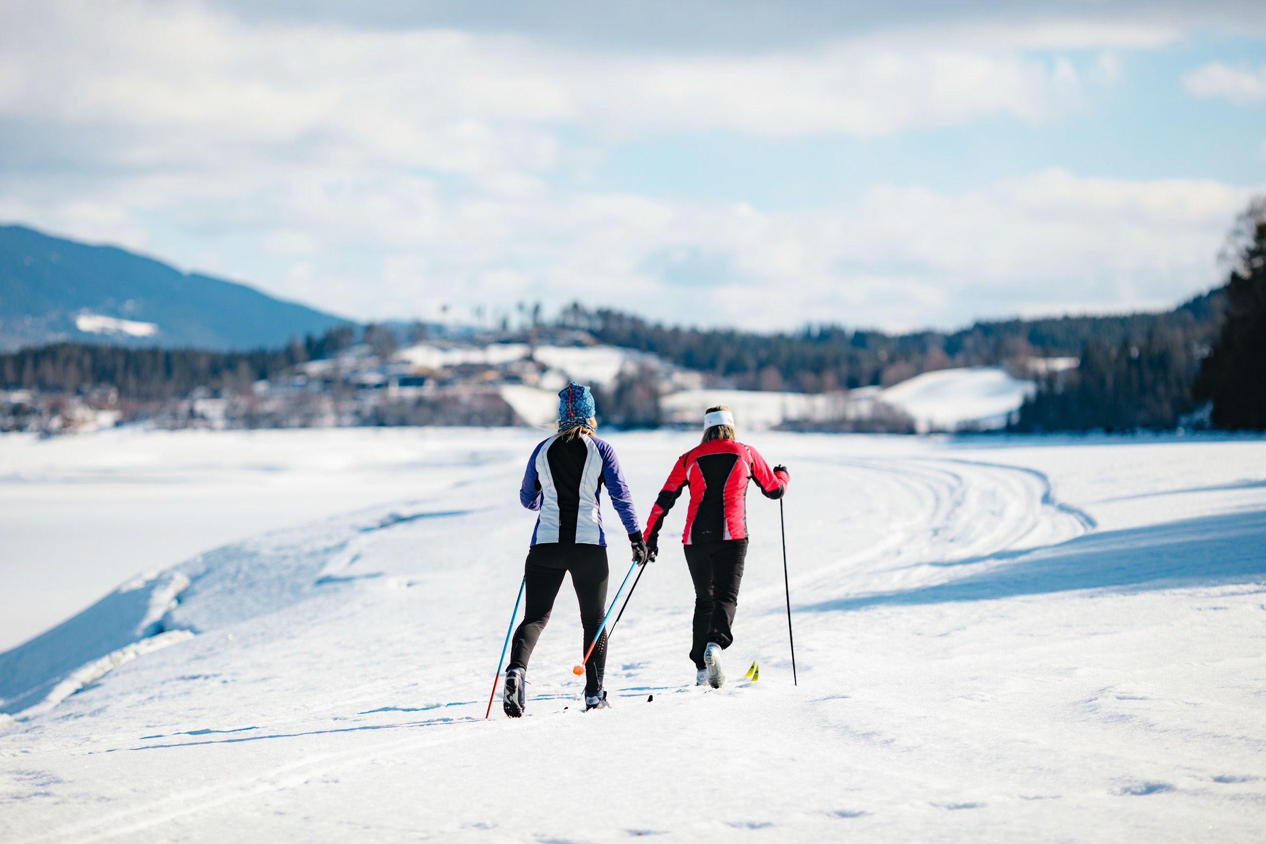Two people cross-country skiing on a snowy path with a mountainous backdrop.