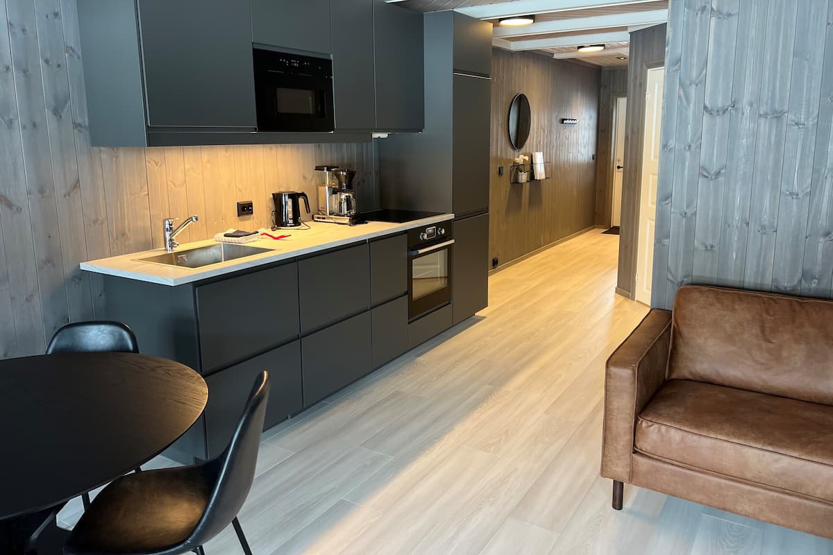 A modern kitchen with dark cabinets, a wooden countertop, and a built-in oven. There is a dining area with a table and chairs, and a brown leather sofa.