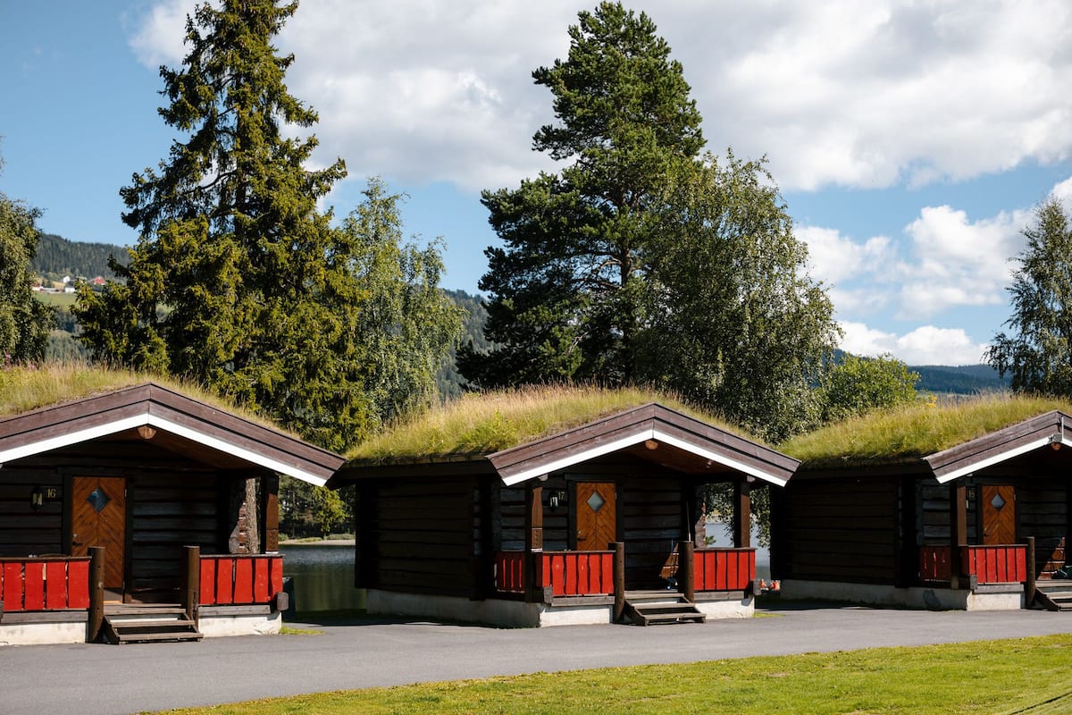 Three log cabins with red railings and wooden doors, set against a backdrop of trees and a clear sky.