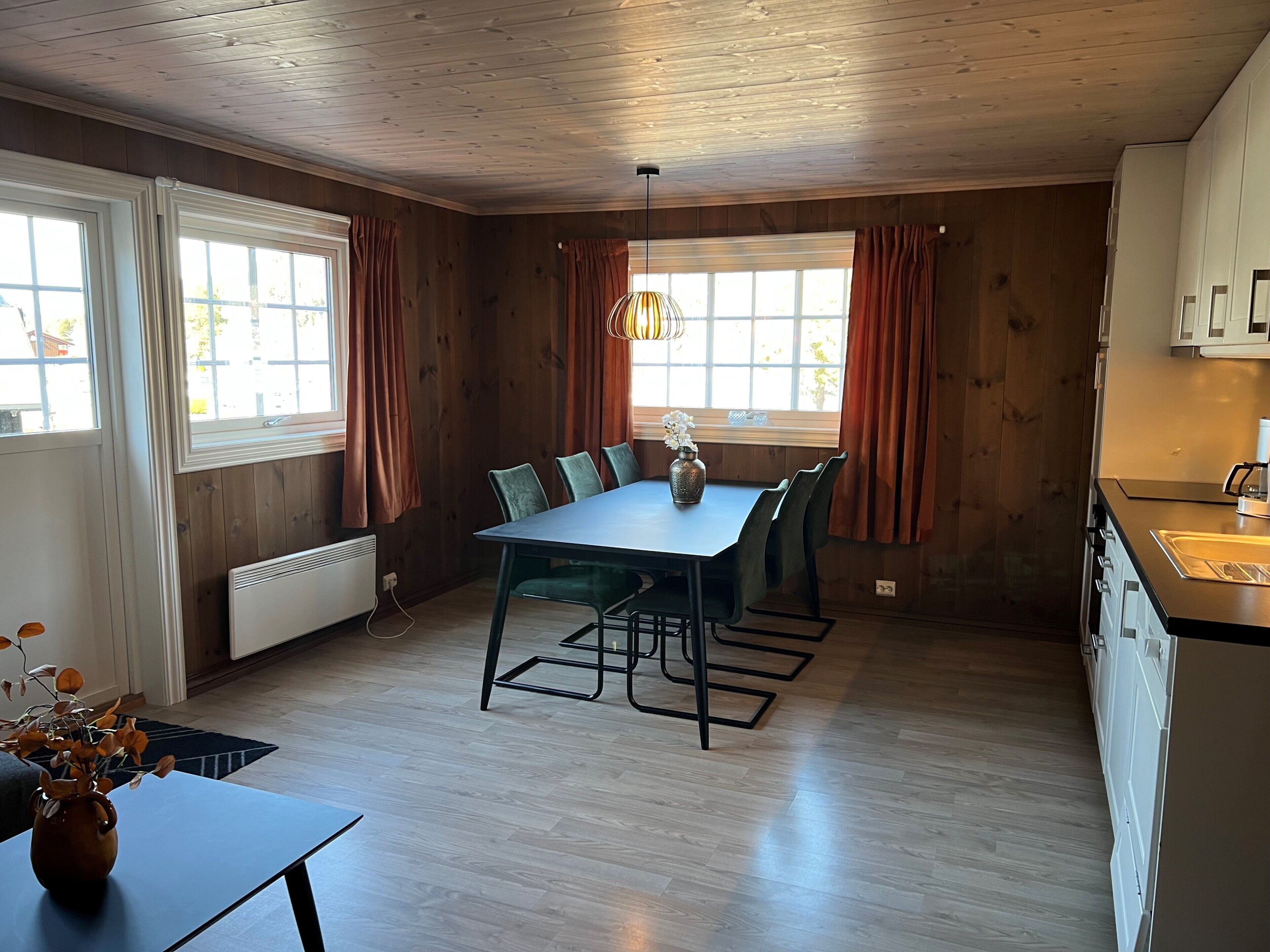 A room with a dining table, chairs, a vase with flowers, a hanging light fixture, and a window with curtains.