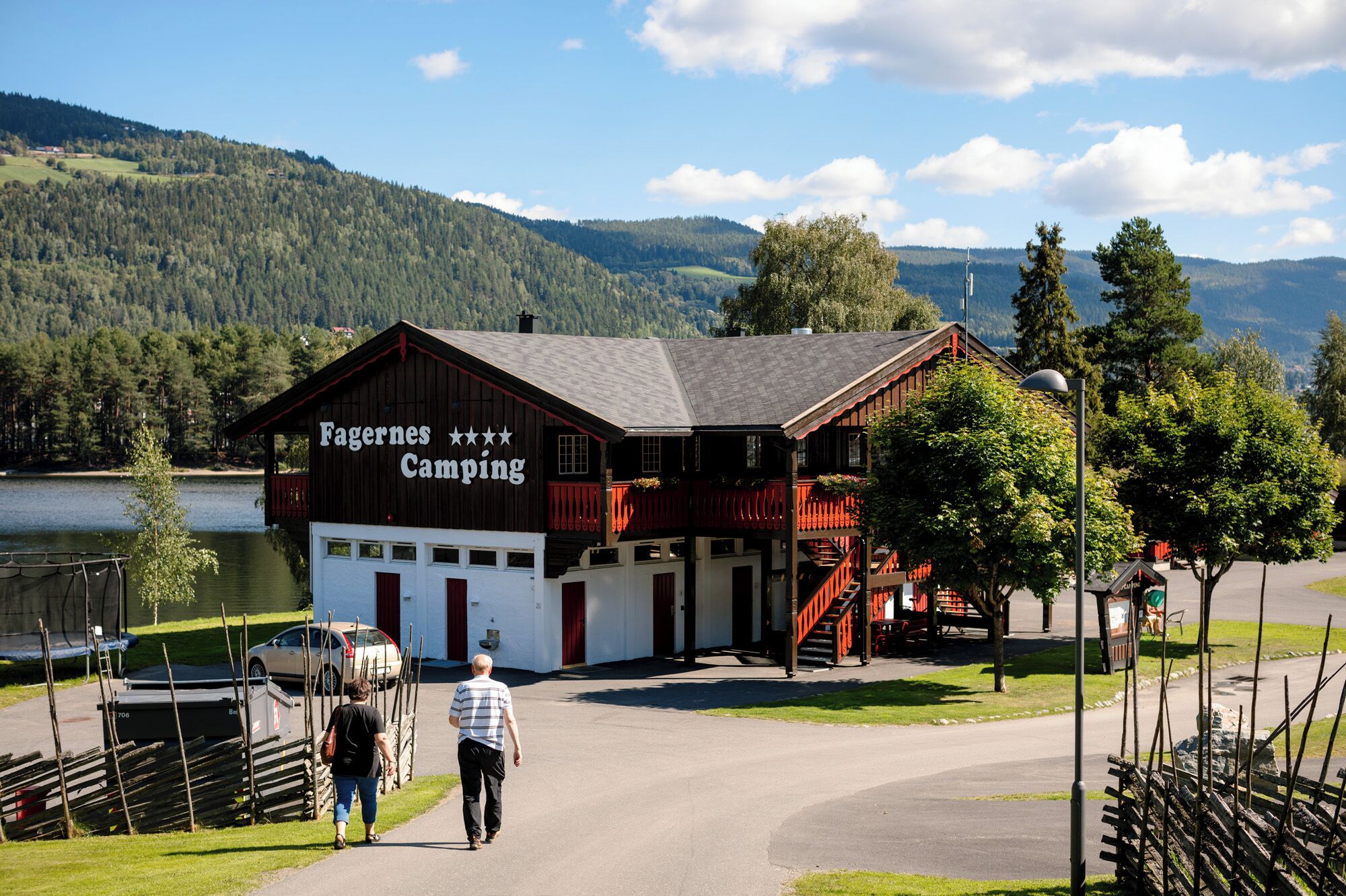 Ein holzhausartiges Gebäude mit einem Schild, auf dem 'Fagernes Camping' steht, und im Hintergrund ein See und Berge.